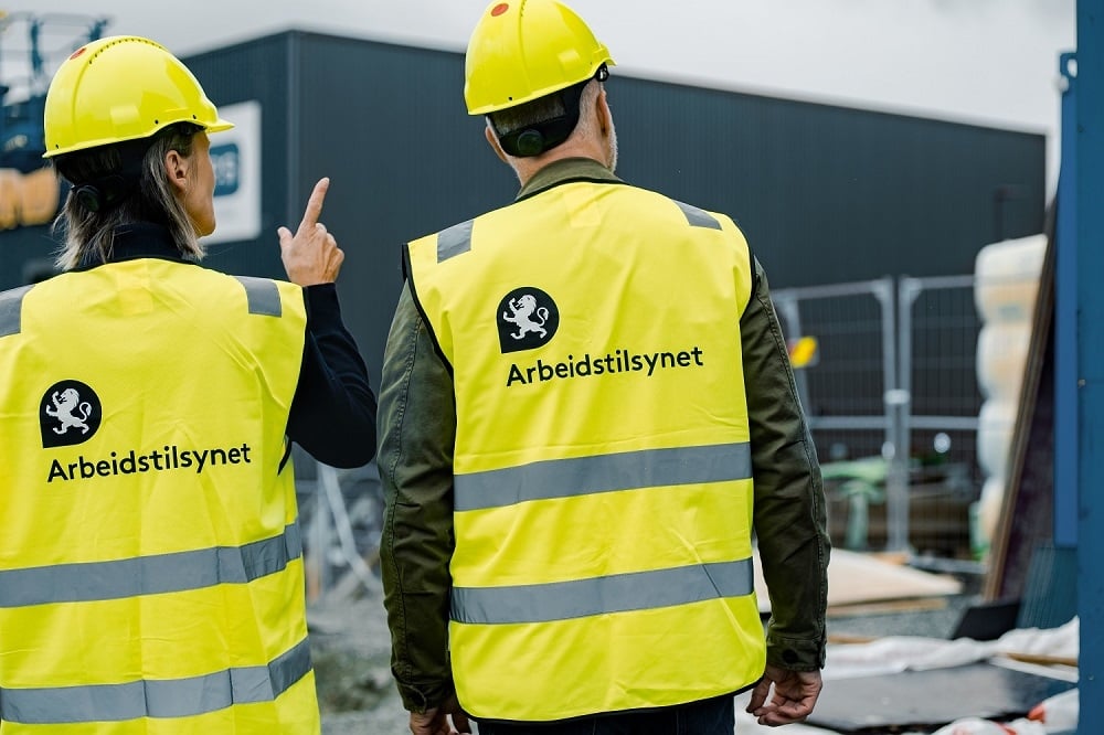 En mann og en kvinne står med ryggen til, de har på seg hjelm og vest med logoen til Arbeidstilsynet. Kvinnen peker på en byggeplass