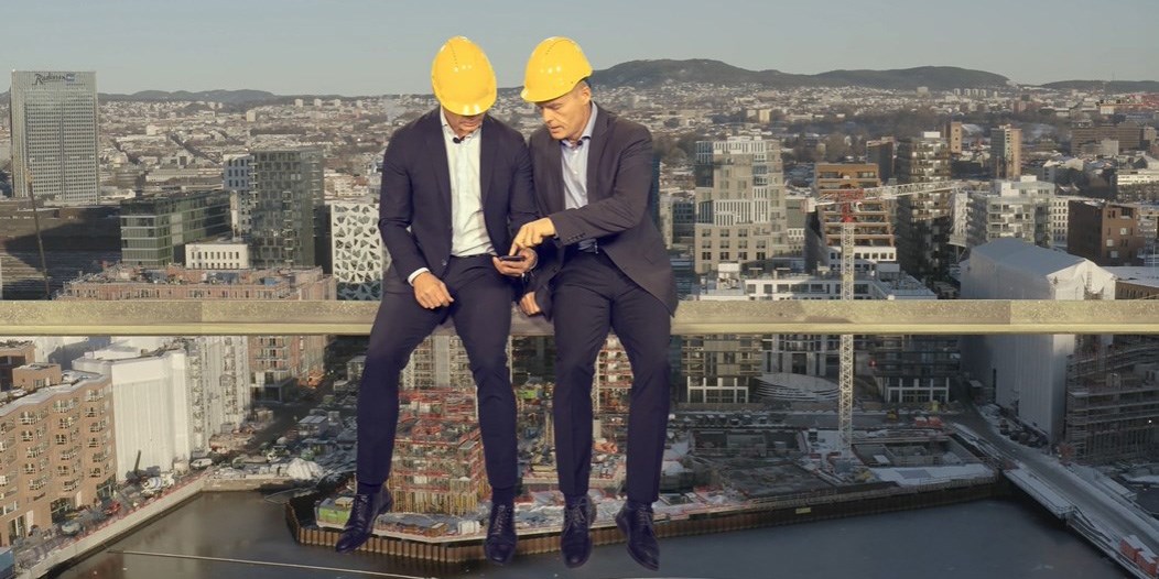 to menn i dress med arbeidshjelm sitter på en bjelke med Oslo i bakgrunnen og titter på en mobiltelefon.